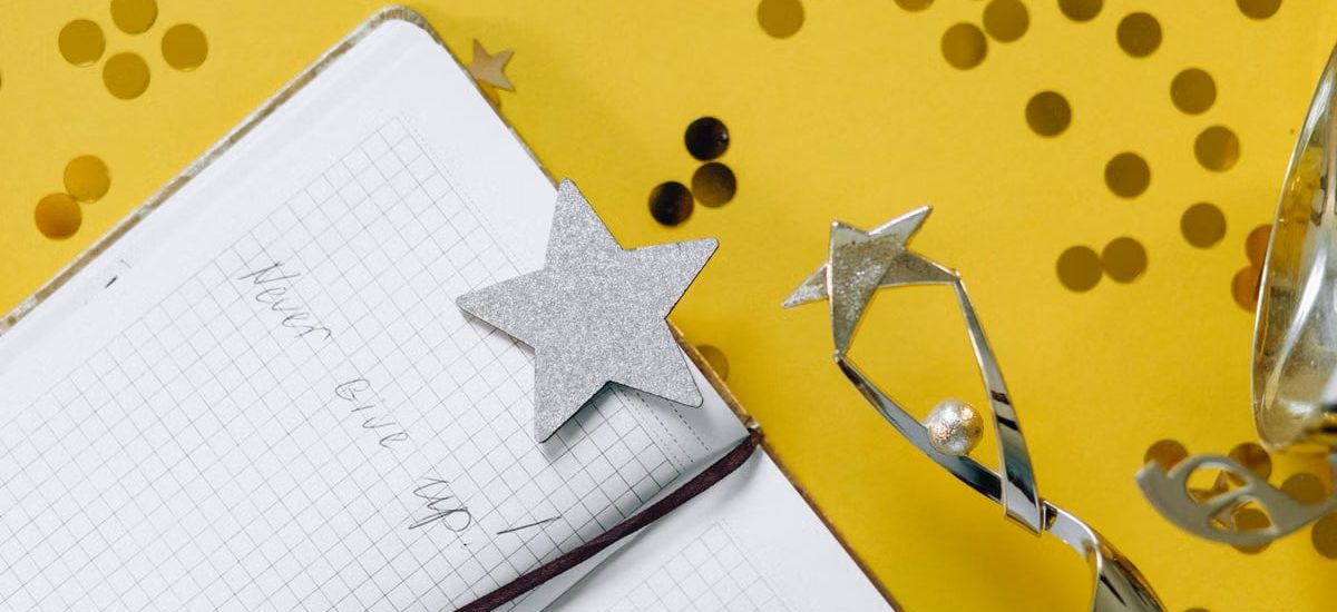 Flat lay of a notebook with motivational text, trophy, and star confetti on a yellow background.